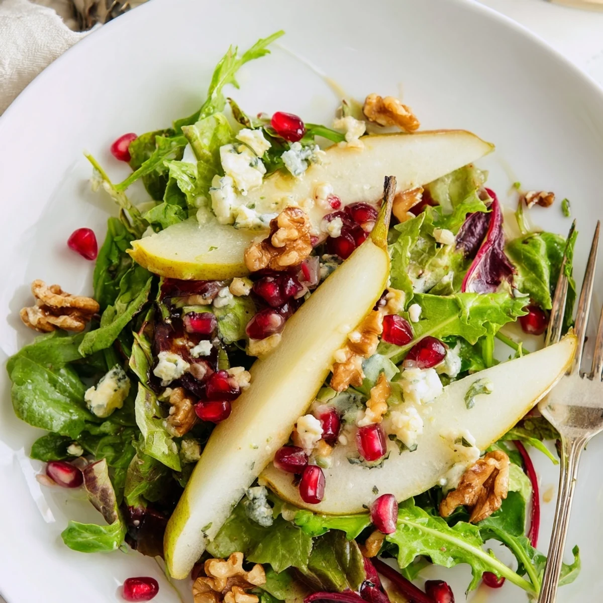 A close-up of the Pear Winter Greens salad featuring fresh baby kale and arugula, garnished with pomegranate seeds and a light honey-Dijon dressing.