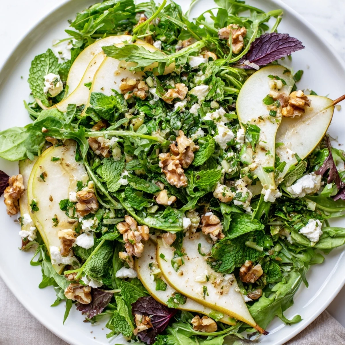 A vibrant bowl of Herbed Pear Greens salad, featuring crunchy walnuts and creamy goat cheese crumbles on a rustic table.