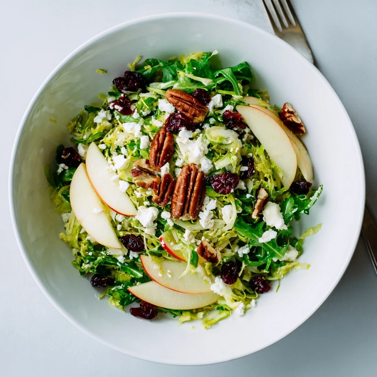 A serving of Shaved Brussels Sprout and Apple Salad Bowl with Candied Pecans garnished with feta and dried cranberries.