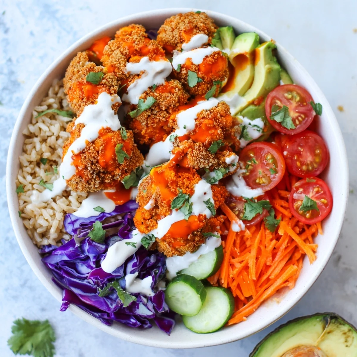Crispy Buffalo Cauliflower Veggie Bowl with Ranch Drizzle is plated over brown rice with fresh avocado and purple cabbage.
