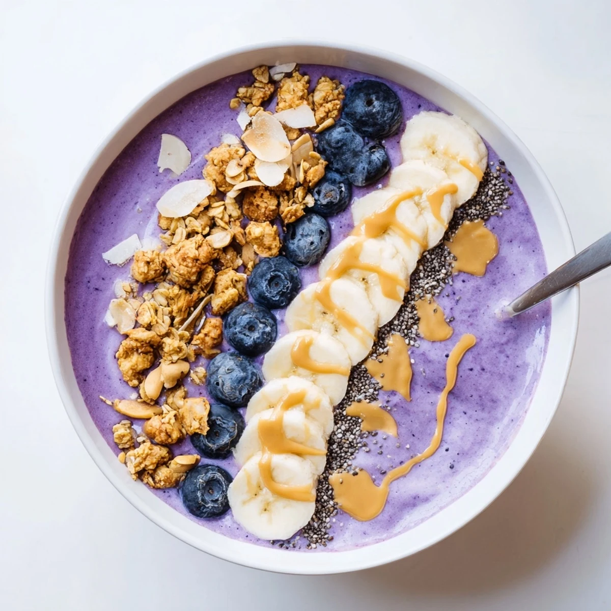 Thick purple smoothie bowl garnished with vanilla cashew cream, toasted granola clusters, and sliced bananas