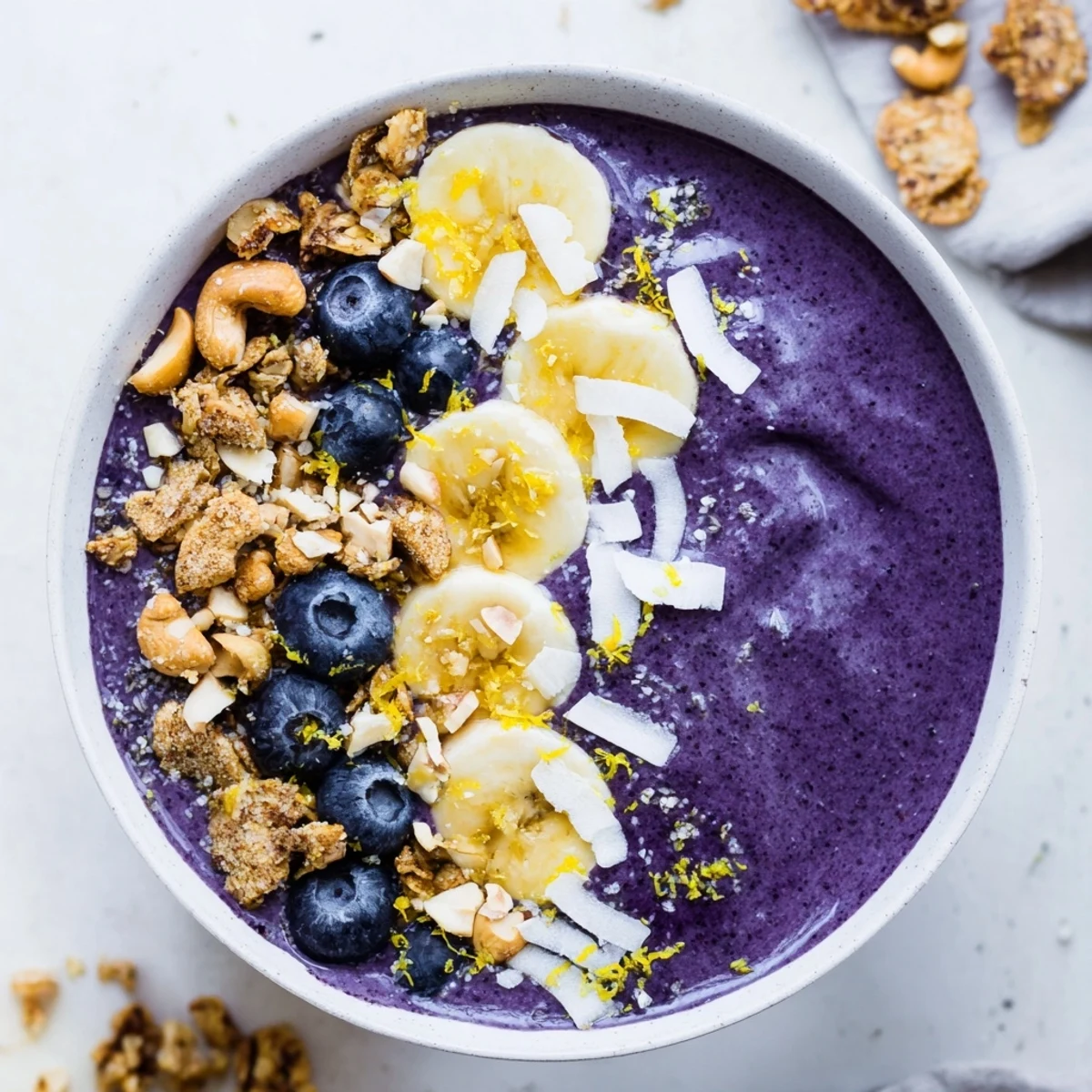 Thick purple blueberry lemon cashew smoothie bowl topped with crunchy golden granola and fresh berries
