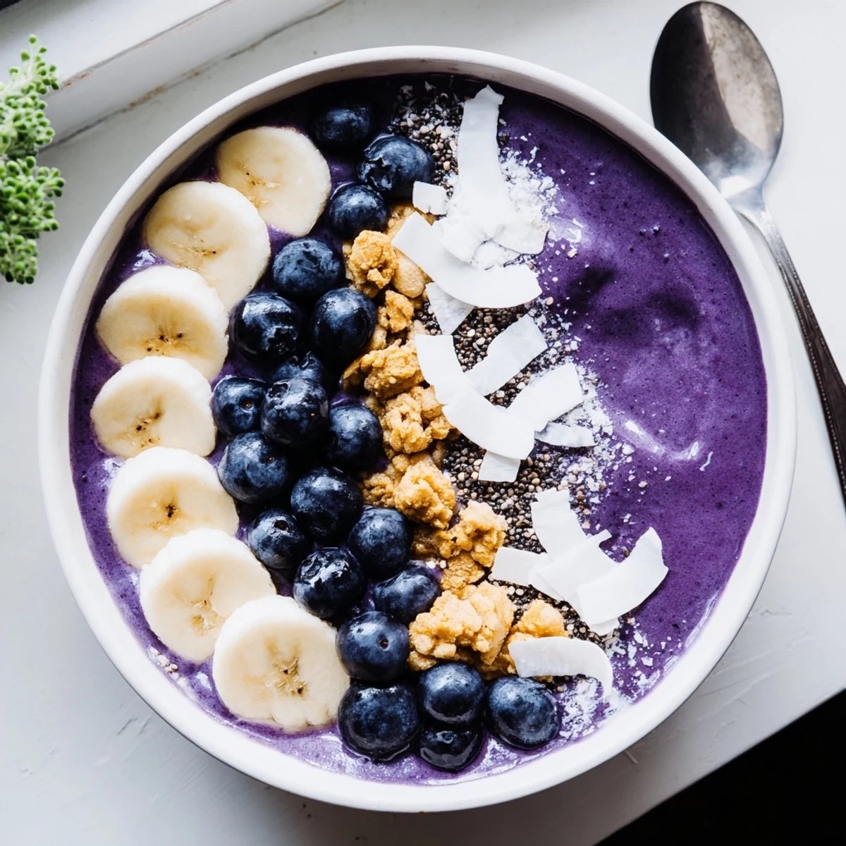 Vibrant purple blueberry banana cashew smoothie bowl garnished with sliced banana and granola