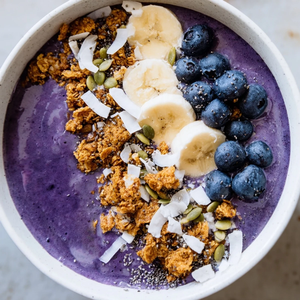 Vibrant blueberry banana coconut smoothie bowl garnished with sliced banana seeds and crispy granola clusters