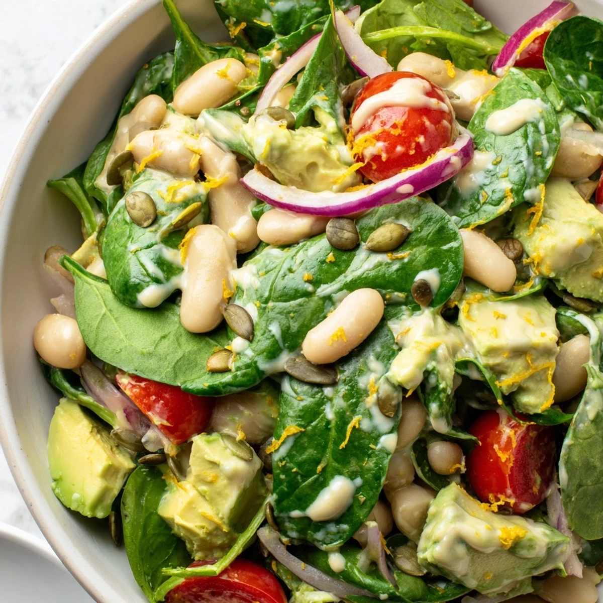 Creamy avocado white bean spinach salad bowl topped with golden pumpkin seeds and lemon zest