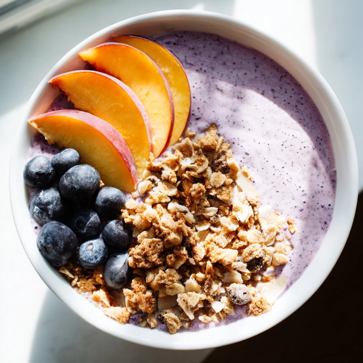 Vibrant blueberry peach almond smoothie bowl topped with golden granola clusters and fresh sliced fruit.
