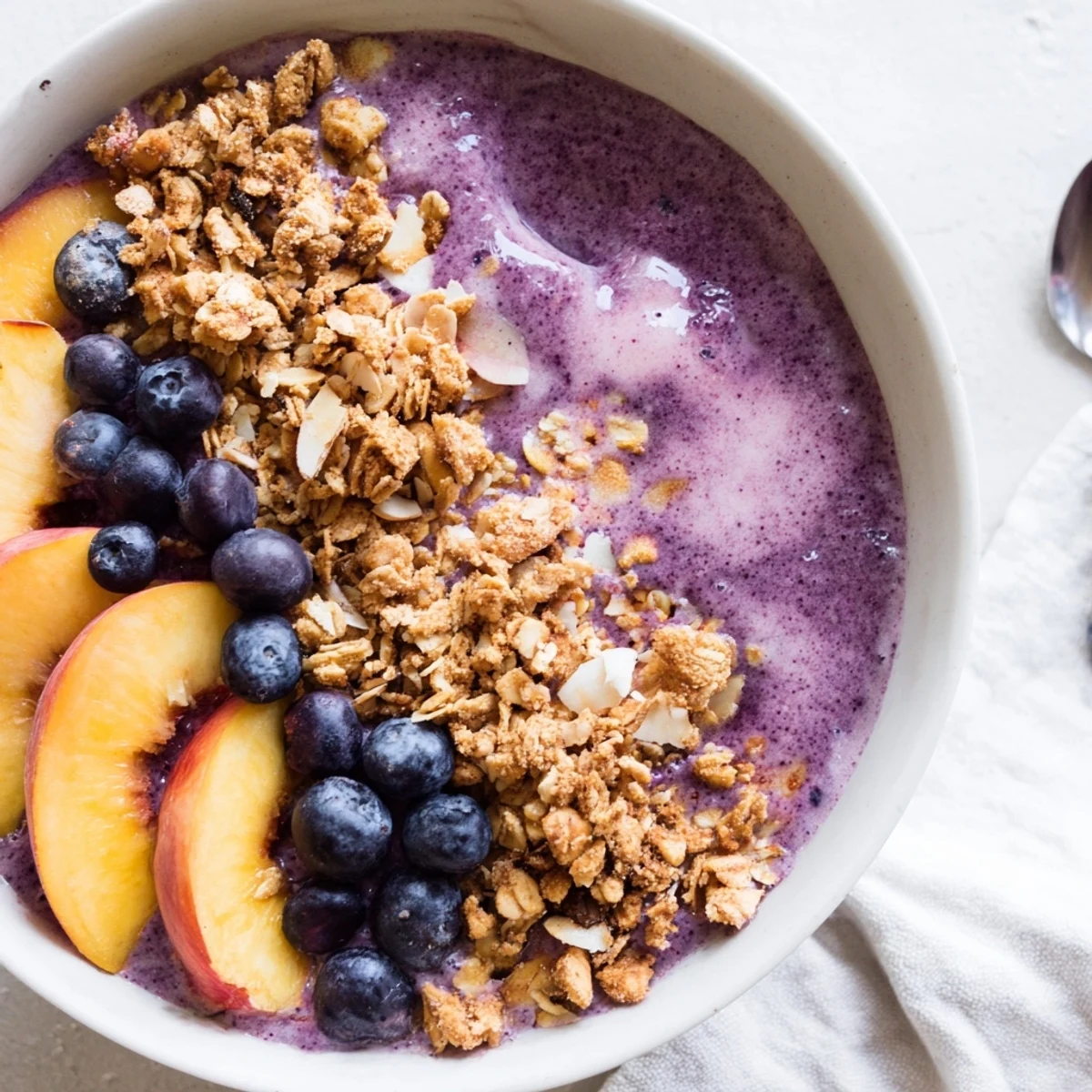 Thick purple smoothie base layered with fresh peaches, blueberries, and crunchy almond granola topping.