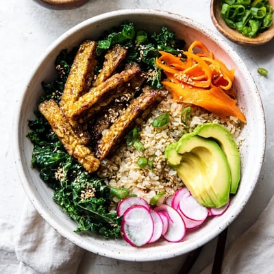 Sliced avocado, julienned carrots, and scallions garnish a vibrant Maple Miso Tempeh Vegan Bowl with sesame greens. 